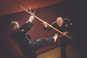 two men in black karate uniforms are fighting with stick against Sai, kobudo weapons