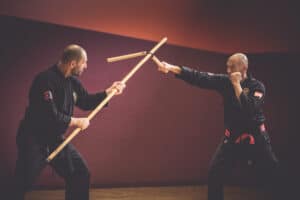 two men in black karate uniforms are fighting with stick against nunchaku, kobudo weapons